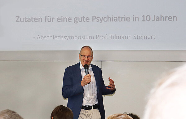 Ein Mann mit Mikrofon in der Hand steht vor einem Menschen in einem Hörsaal.