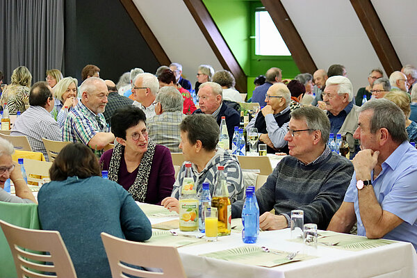 In einem Festsaal sitzen an mehreren festlich gedeckten Tischreihen ältere Damen und Herren beisammen und unterhalten sich miteinander.