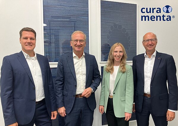 Drei Männer in Anzügen und eine blonde Frau mit hellgrünem Jacket stehen nebeneinander. 