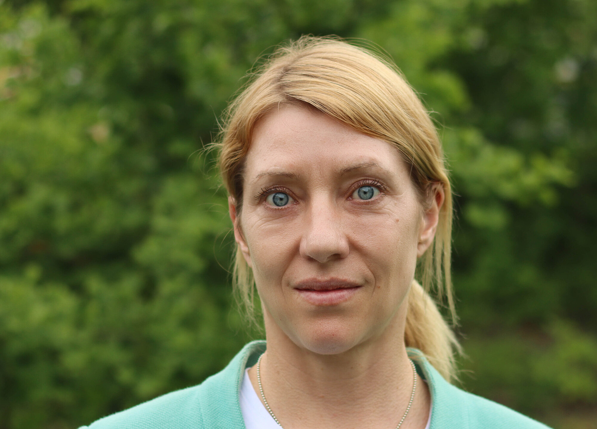 Porträtfoto einer Frau vor grauem Hintergrund. Die Frau hat lange blonde glatte Haare mit Seitenscheitel zu einem Zopf gebunden. Sie trägt ein blaues Oberteil mit heller Weste darüber. Sie trägt große weiße runde Ohrhänger. Sie hat blaue Augen und schaut freundlich in die Kamera.