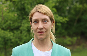 Porträtfoto einer Frau vor grauem Hintergrund. Die Frau hat lange blonde glatte Haare mit Seitenscheitel zu einem Zopf gebunden. Sie trägt ein blaues Oberteil mit heller Weste darüber. Sie trägt große weiße runde Ohrhänger. Sie hat blaue Augen und schaut freundlich in die Kamera.