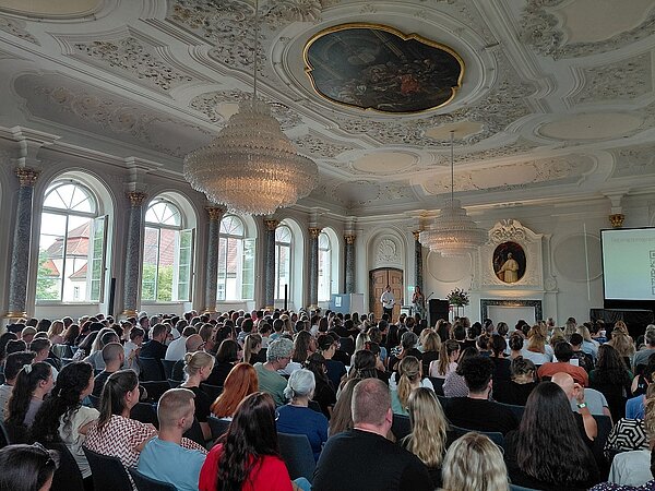 Zu sehen ist ein vollbesetzter, bestuhlter Klostersaal, fotografiert von hinten. Die Sitzenden blicken auf eine kleine Bühne nach vorne, wo zwei Personen moderieren.