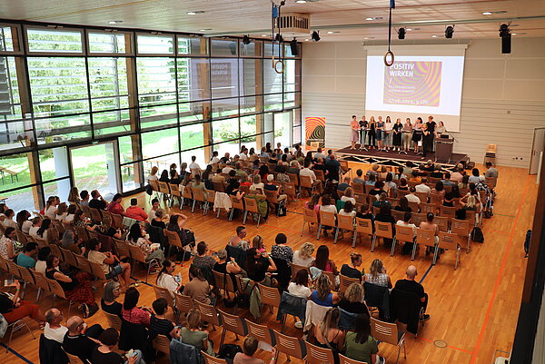 Blick von oben auf eine vollbesetzte Halle, auf der Bühne stehen mehrere referierende Personen.