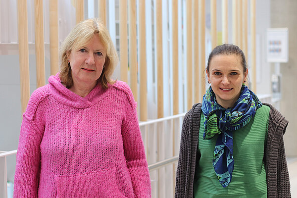 Eine blonde und eine brünette Frau stehen lächelnd in einem hellen Flur.