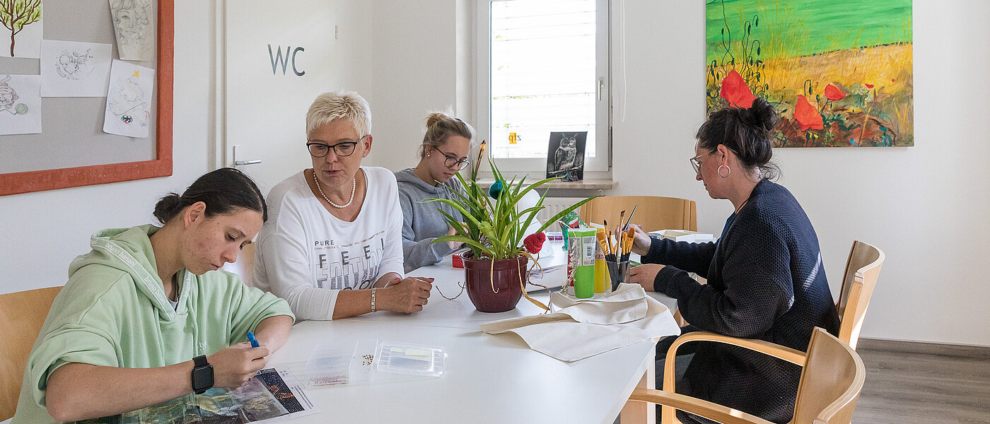 Vier Frauen unterschiedlichen Alters sitzen in einem hellen Raum um einen großen Tisch und führen unterschiedliche Bastelarbeiten aus, eine der Frauen leitet die anderen dabei an. 