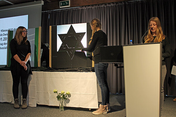 Eine Frau steht an einem Pult, zwei weitere Frauen stehen links im Bild.