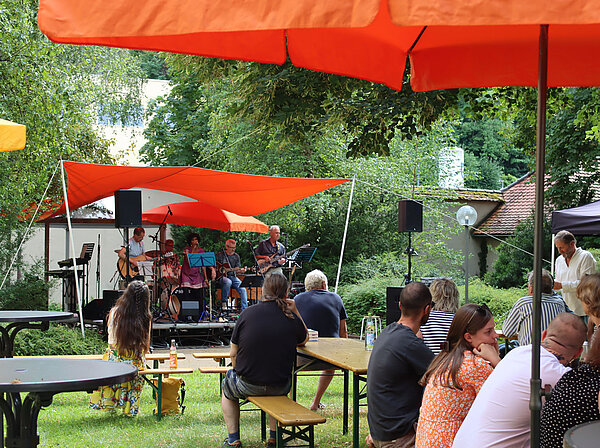 Menschen sitzen auf Bierbänken unter bunten Sonnenschirmen in einem Park, an dessen Ende eine Bühne mit Musikern zu sehen ist. 