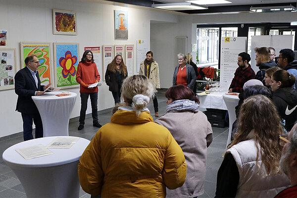 In einem großen hellen Raum hängen Bilder und Infotafeln an den weißen Wänden, das Foto entstand bei der Eröffnung der Ausstellung, im Hintergrund stehen die Redner:innen, im Vordergrund interessierte Zuhörer:innen.