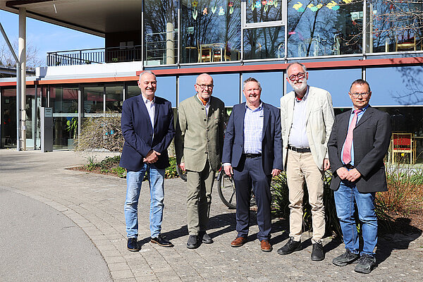 Fünf Männer in Business-Kleidung stehen vor dem Eingang eines Heimgebäudes in der Sonne und lächeln in die Kamera.