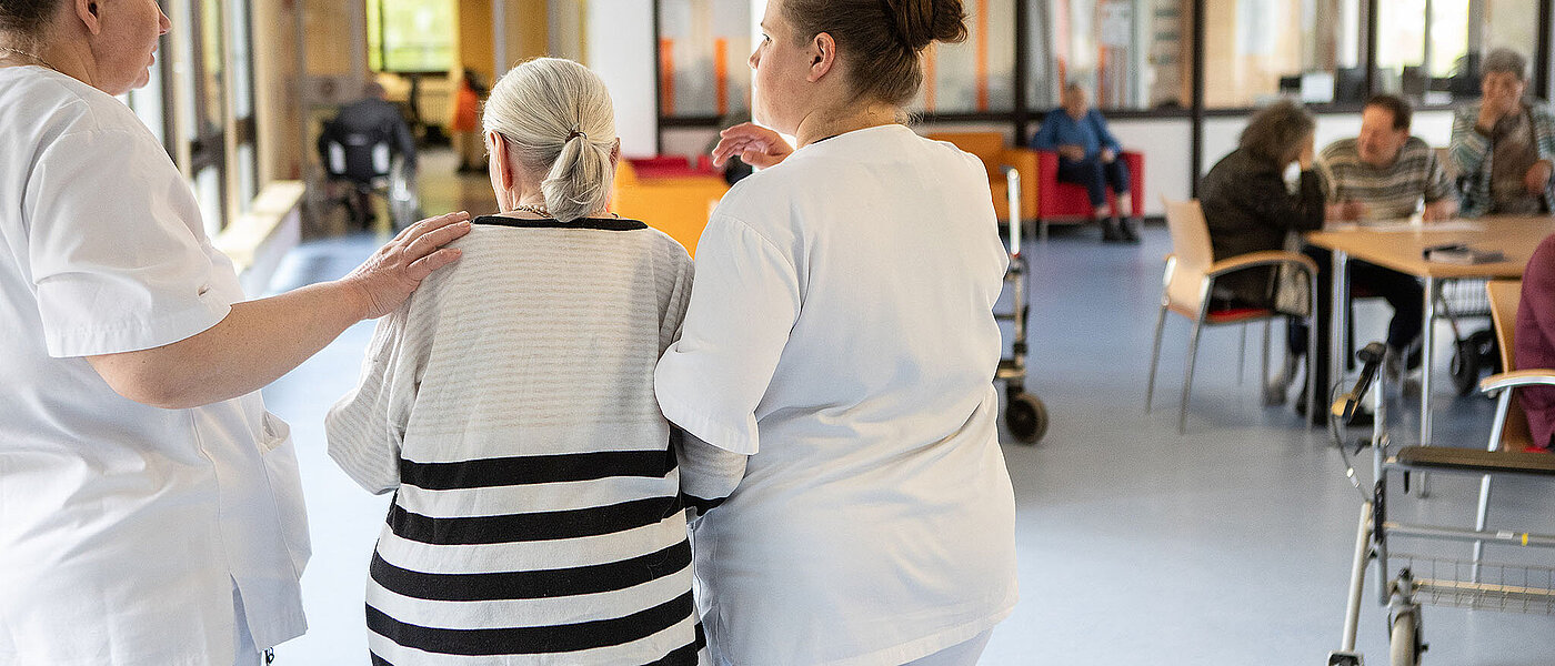 Eine junge Pflegerin unterstützt eine ältere Dame im blau gestreiften Pullover beim Gehen
