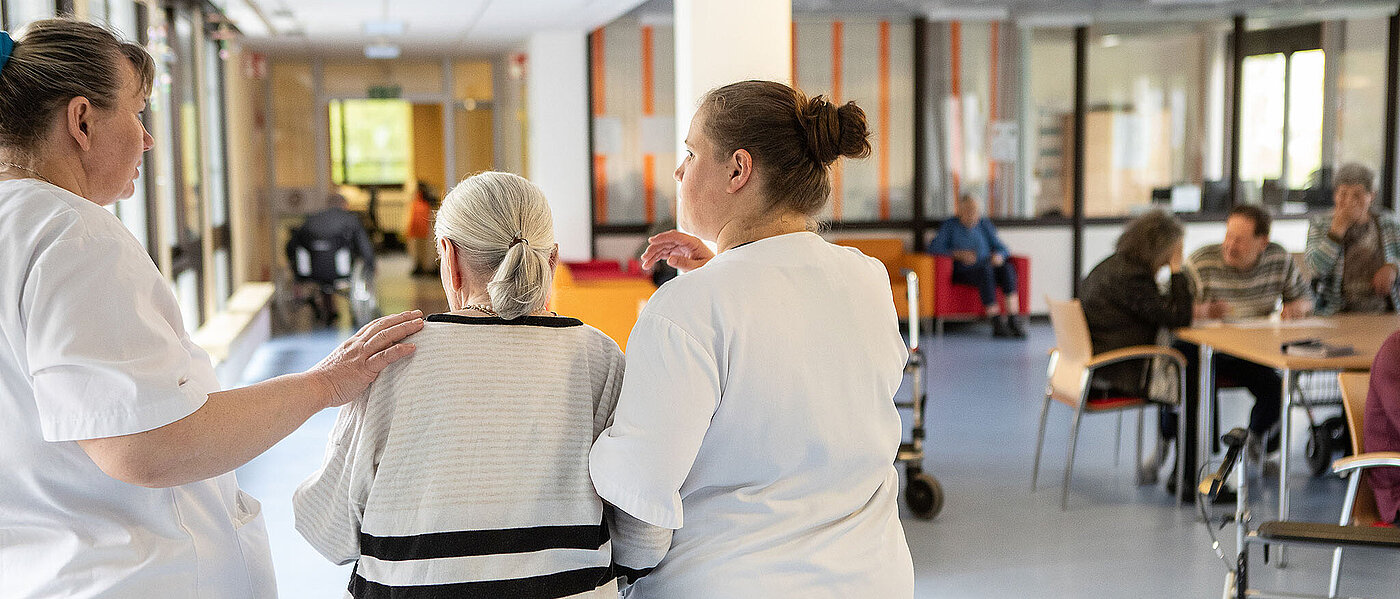 Eine junge Pflegerin unterstützt eine ältere Dame im blau gestreiften Pullover beim Gehen