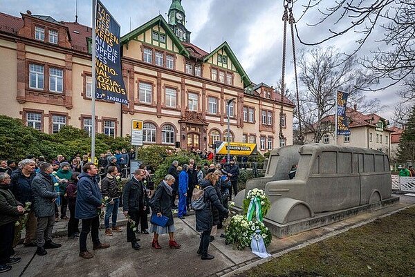 Man sieht eine große Menschengruppe, im Vordergrund ist ein Bus aus grauem Beton, im Hintergrund historische Fachwerkgebäude.  