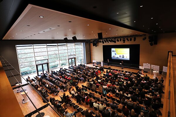 In einem großen Saal sitzen 350 Teilnehmende auf Stühlen, auf der Bühne stehen zwei Personen, die etwas erklären. 