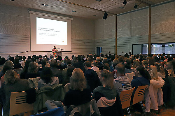 Eine vollbesetzte, bestuhlte Turnhalle. Die Zuhörer:innen blicken auf eine Frau mittleren Alters, die einen Vortrag hält. Hinter ihr an der Wand ist eine PowerPoint-Präsentation zu sehen.