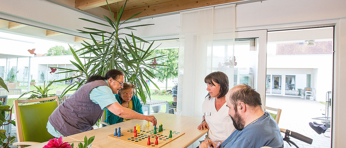 Eine Gruppe Menschen sitzt um einen großen Holztisch und spielt Mensch-Ärgere-Dich-Nicht. Eine Frau trägt Pflegekleidung, die restlichen Personen sind alltäglich gekleidet. 