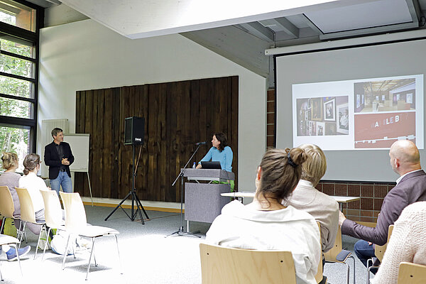 Moderator und Referentin tauschen sich aus, Zuhörerinnen und Zuhörer im Vordergrund, im Hintergrund rechts ist eine Bildschirmpräsentation zu sehen.