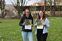 Zwei Frauen stehen nebeneinander auf einer grünen Wiese, am oberen Bildrand ist ein Banner "yes we care" zu sehen. 