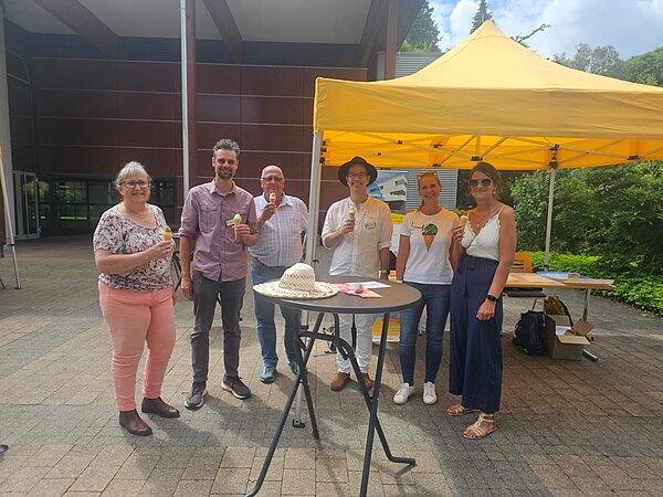 Eine Gruppe Menschen steht vor einem gelben Pavillon, alle halten ein Eis in der Hand. 