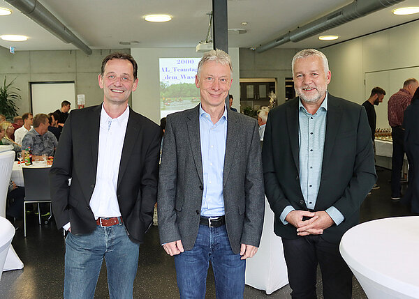 Drei Herren, gekleidet mit Hemd und Jacket, lächeln in die Kamera. Im Hintergrund ist ein mit Menschen gefüllter Saal zu erkennen.