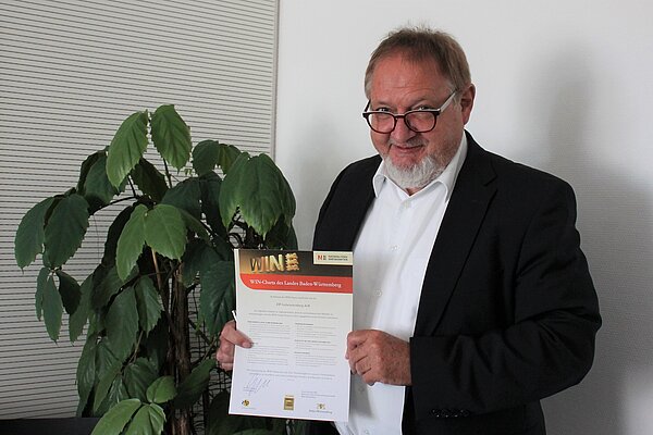 Ein Mann steht vor einer Pflanze und zeigt stolz ein Dokument in die Kamera. Der Mann trägt ein schwarzes Sakko, ein weißes Hemd darunter und eine Brille. Er hat einen grauen Bart.