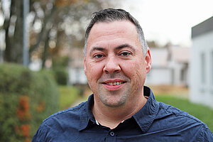 Ein Mann mittleren Alters mit kurzen braunen Haaren und dunkelblauem Hemd steht lächelnd auf einem Weg im Freien.