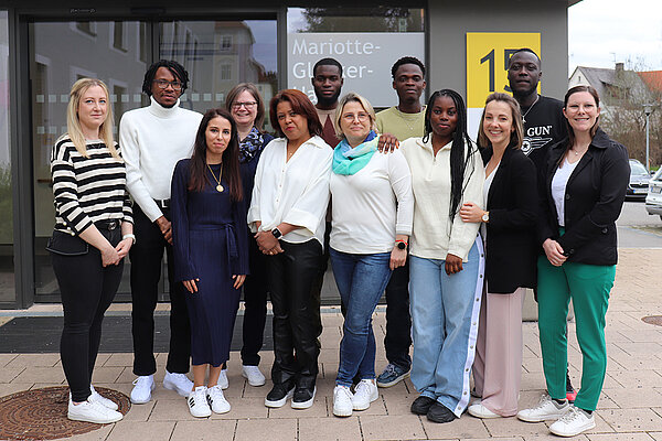 Eine Gruppe junger Menschen aus verschiedenen Herkunftsländern steht vor einem Gebäude, welches mit einem gelben Schild mit der Nummer 15 gekennzeichnet ist.