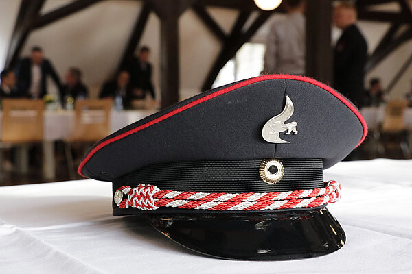 Ein Feuerwehrhut liegt auf einer weißen Tischdecke, im Hintergrund sind Feuerwehrleute sitzend und stehend im Gespräch.
