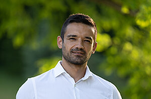 Ein dunkelhaariger Mann mit Dreitagebart und weißem Hemd vor einem grünen Hintergrund.