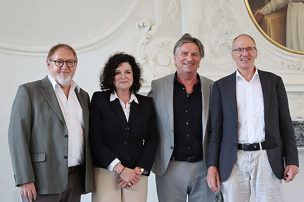 Drei Männer und eine Frau stehen nebeneinander, sie sind alle förmlich gekleidet und lächeln. 