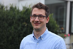 Porträtfoto eines jungen Mannes. Er hat kurze dunkelbraune Haare, trägt eine eckige Brille mit dunklem Rahmen und ein hellblaues Hemd mit Kragen. Er lächelt mit geschlossenem Mund in die Kamera. Im Hintergrund sind verschwommen ein Gebäude und ein Busch zu sehen.