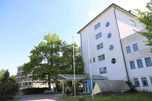 Man sieht ein mehrstöckiges helles Gebäude mit einem überdachten Eingang. 