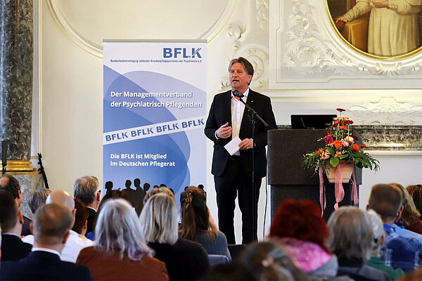 Ein Mann in weißem Hemd und schwarzem Anzug steht in einem historischen Festsaal mit stuckverzierten Wänden vor zahlreichen Zuhöherinnen und Zuhörern.