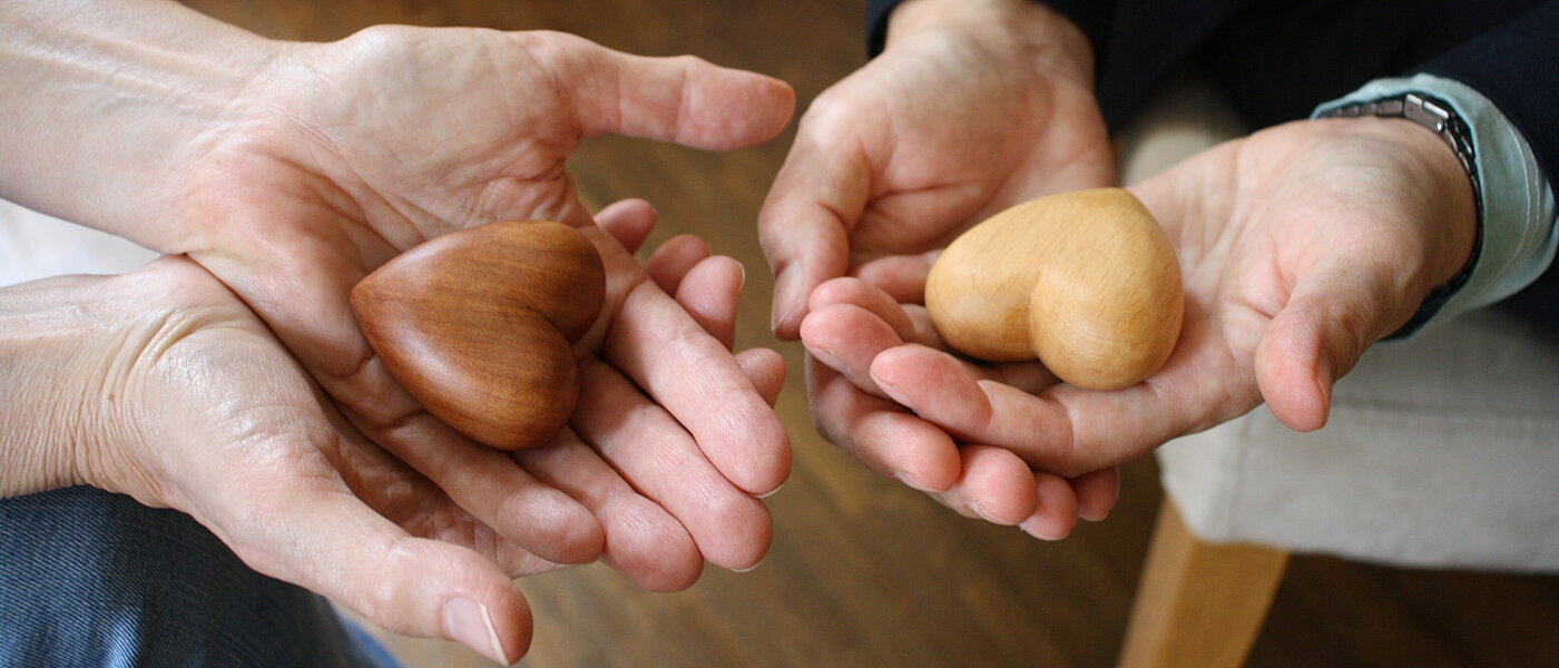 Man sieht die geöffneten Hände zweier Menschen, in die Händen liegt ein kleines Herz aus Holz. 
