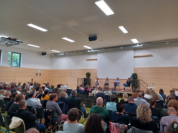 In einem großen Saal sitzen viele Menschen nebeneinander auf Stühlen und blicken konzentriert zur Bühne, auf der vier Frauen nebeneinander sitzen und vortragen. 
