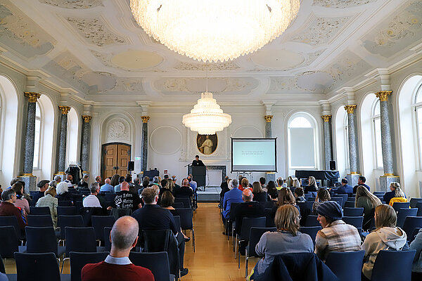 Blick in einen barocken Festssaal mit Holzparkett, Stuck und Kronleuchtern, in dem zahlreiche Zuhörer:innen auf Stuhlreihen einem Redner am Rednerpult gegenüber sitzen.