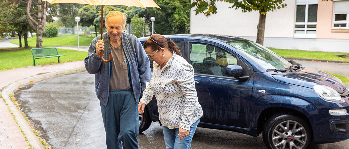 Eine Frau und ein Mann laufen gemeinsam unter einem gelben Regenschirm von einem Auto weg.