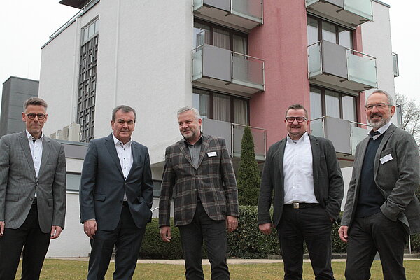 Vor einem hohen Gebäude mit Balkonen stehen fünf ältere Männer, die alle ein Sakko tragen.