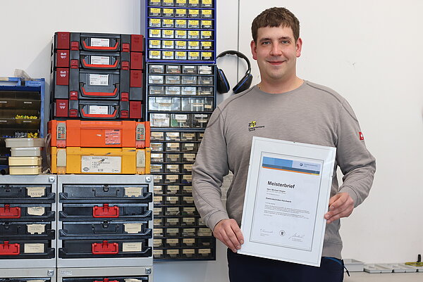 Ein Mann steht neben bunten Werkzeugkoffern und hält seinen Meisterbrief in der Hand. Er lächelt sanft in die Kamera.