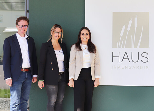 Ein Mann und zwei Frauen stehen vor einem Schild mit der Aufschrift "Haus Irmengardis". 