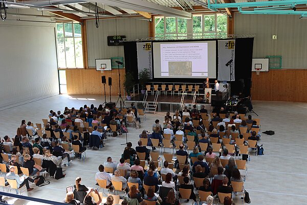 Eine Halle aus der Vogelperspektive, es sitzen Menschen auf Stuhlreihen vor einer Bühne. 