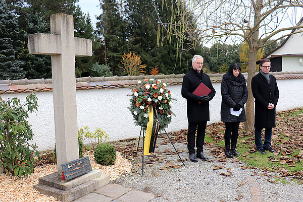 Drei Personen stehen würdevoll neben einem Mahnmal, ein Kranz wurde niedergelegt.