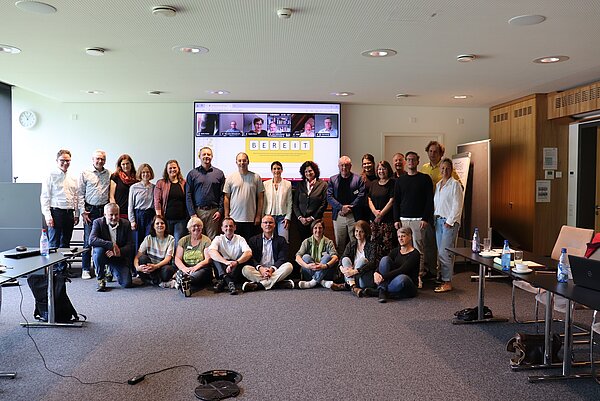 Eine große Gruppe Menschen gruppiert sich vor einem Bildschirm, auf dem Bildschirm sind die Teilnehmenden einer Videokonferenz zu sehen. 