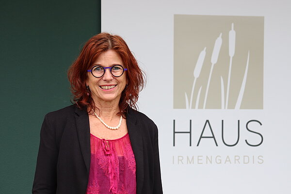 Eine Frau mit roten Haaren und Brille steht vor einer dunkelgrünen Wand. 