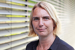Portraitbild Andrea Sonntag, eine Frau mit Seitenscheitel, kurzen blonden Haaren zum Kinn. Sie steht vor einer Jalousie.