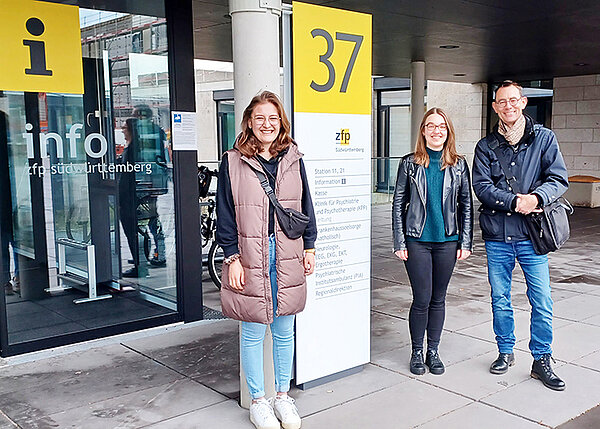 Drei Personen stehen vor einer weiß-gelben Infostele.
