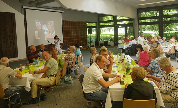 Eine Gruppe von Pensionären sitzen an Tischen in einer großen Hallo, vorne spricht eine Frau ins Mikrofon.