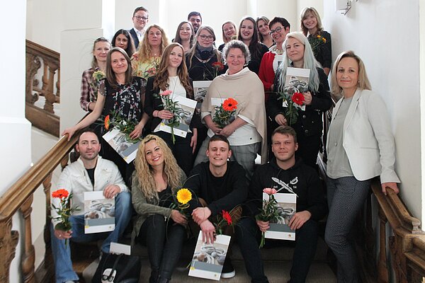 Absolventen der Weiterbildung sind auf einer Treppe platziert. Jeder hält sein Abschluss-Dokument in der Hand und eine Blume in gelb oder rot.