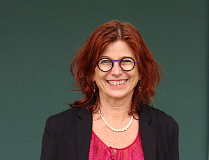 Eine Frau mit roten, halblangen Haaren und Brille die eine Perlenkette, einen schwarzen Blazer und eine pinke Bluse trägt.