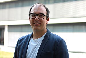 Porträtfoto eines Mannes mit dunklen kurzen Haaren und eckiger dunkler Brille. Er trägt ein helles Oberteil und einen dunkelblauen Sakko darüber. Er lächelt in die Kamera, im Hintergrund ist verschwommen ein Gebäude zu sehen.