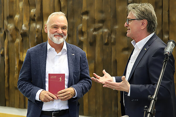 Zwei Männer in Business-Anzügen stehen vor einer holzgetäfelten Wand, der linke schaut in Richtung Kamera und hat ein Buch in der Hand, der rechte spricht in Richtung des anderen.
